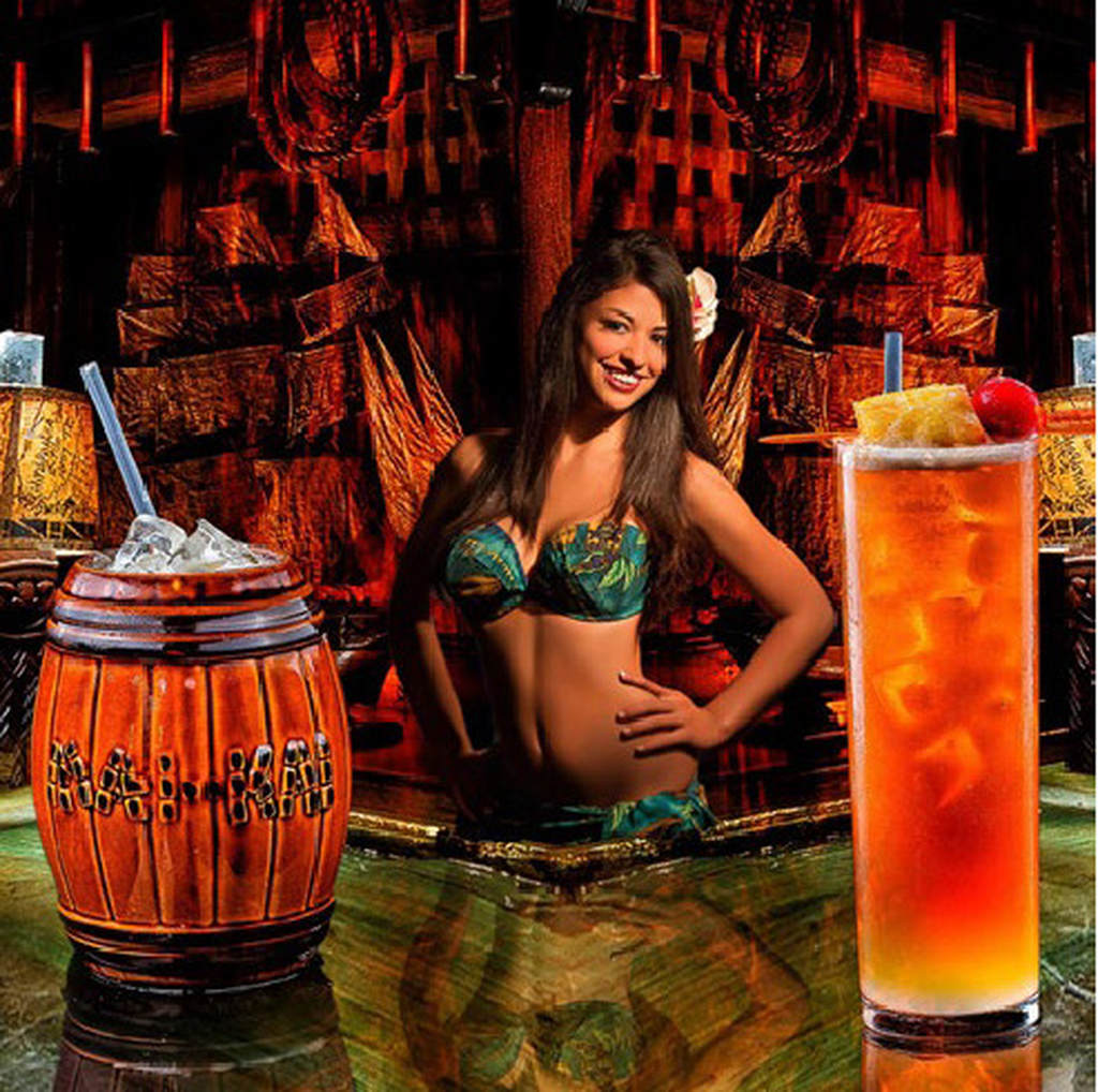 a woman bartender in a bikini posing behind a wooden bar with two drinks in front of her
